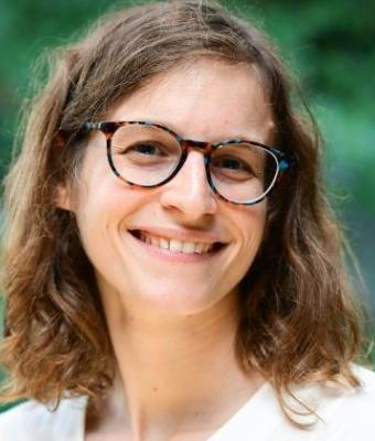 Anna Callis, a smiling woman with curly brown hair and glasses, wearing a white top, outdoors with a blurred green background.