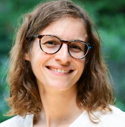 Anna Callis, a smiling woman with curly brown hair and glasses, wearing a white top, outdoors with a blurred green background.