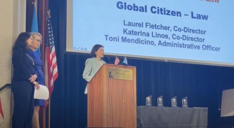 Katerina Linos speaks at the podium during the 19th Annual Global Citizen Awards. Behind her, a screen reads "Global Citizen – Law" with her name, along with Laurel Fletcher, the institute's co-director, and Toni Mendicino, the institute's administrator. Two women stand nearby, with American and UN flags on the podium, U.S. flags in the background, and awards on a table.