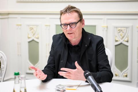 James Robinson speaking at a table during a formal discussion, with a microphone and papers in front of him.